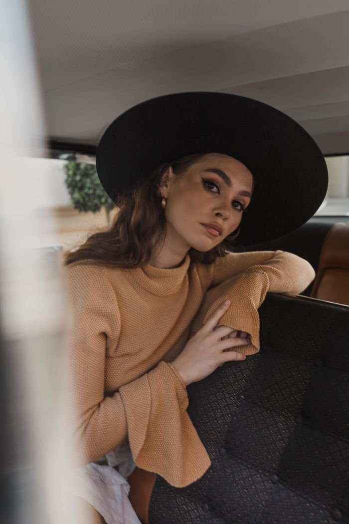 Home Stylish woman posing in car interior wearing a black hat and beige sweater.